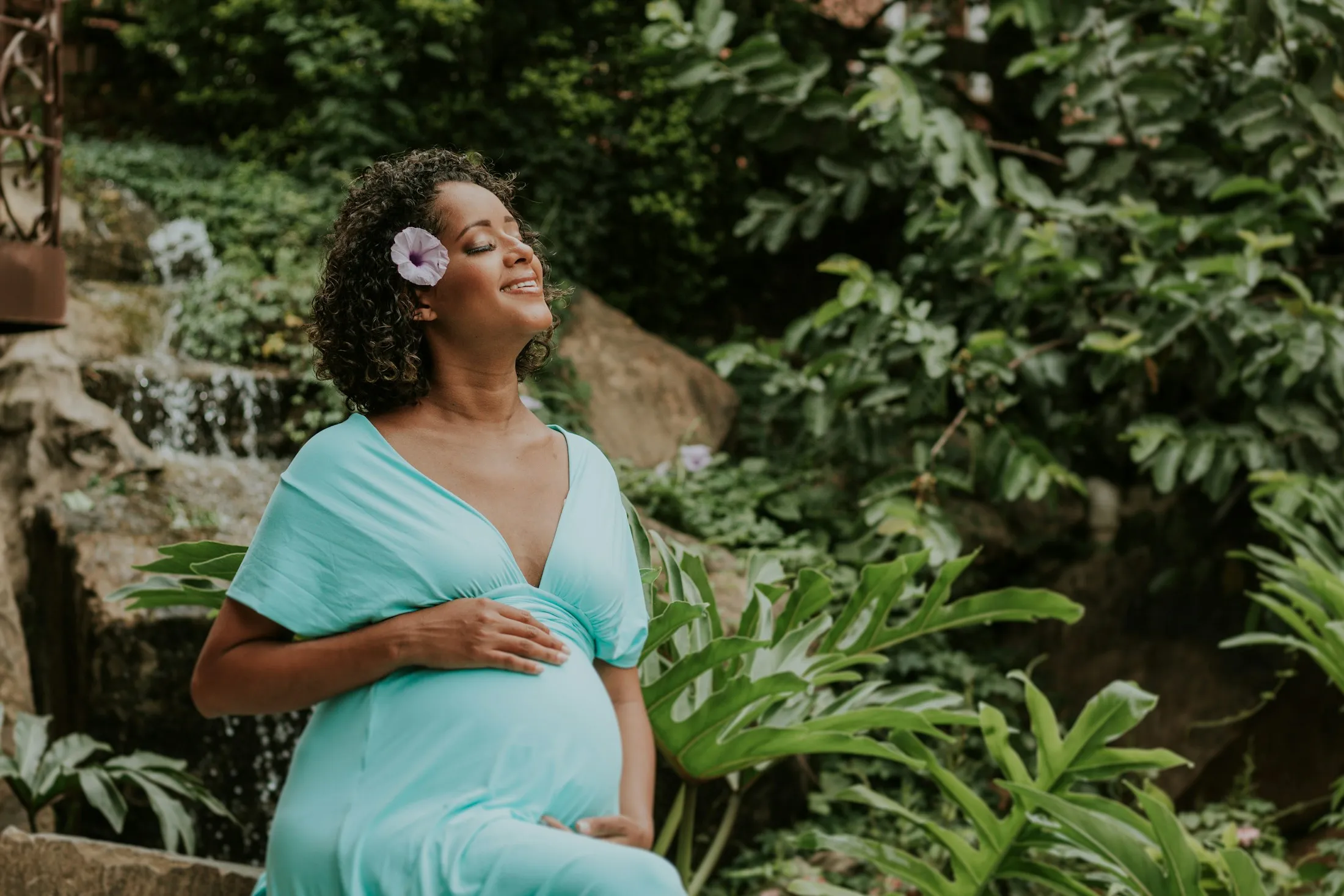A pregnant woman with a flower in her hair, eyes closed, beside a garden waterfall